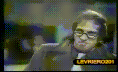 Video gif. A man looks at us with half circle glasses on his face. He has a big grin on his face while he makes an “A-ok” symbol with his hand. 