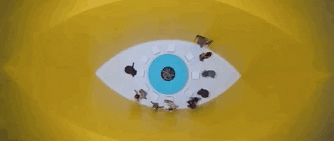 Digital art gif. We look down into a yellow eye shaped room as people circle around a blue pupil on the floor. 
