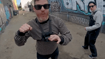 Music video gif. Andy Hurley, the drummer of Fall Out Boy, wearing a striped shirt and sunglasses swings his arm around like he's swinging a punch in an alley, just barely missing the camera. 