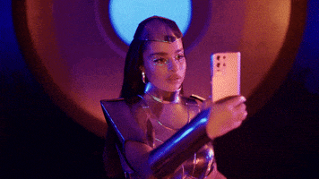 Music video gif. From the video for Thought About That, Noa Kirel is dressed in a metallic futuristic outfit, moving like a cyborg as she twists her torso holding a device, then carefully examines her hand.