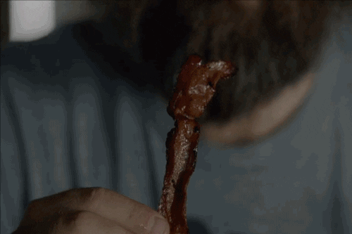 Video gif. We follow the shot of bacon entering a man's mouth as he eats the whole piece in one bite. His eyes roll back in his head at first contact and his bangs blow back slightly as he chews.