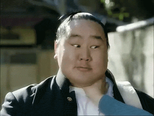 Video gif. Someone has an Asian man in a chokehold, but he gives them a thumbs up and blinks both eyes in a failed attempt to wink.