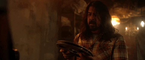 Celebrity gif. Dave Grohl of Foo Fighters holds a dusty film reel in both hands, then bends over and blows the dust off the top.