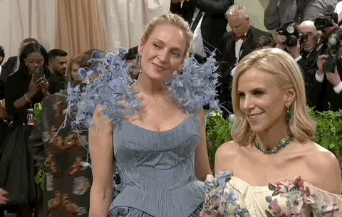 Met Gala 2024 gif. Uma Thurman poses with Tory Burch, wearing one of her designs, a powder blue gown with full pleats, structured bodice, flowers growing tall out of her shoulders.