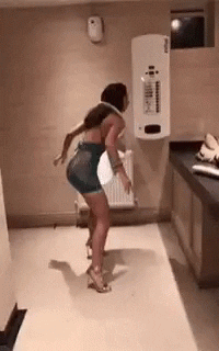 Video gif. A woman standing in a public bathroom has a toilet lid around her neck. She swings it around her neck like a hula hoop and drunkenly stumbles backwards.