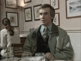 TV gif. Sitting at a restaurant table, Steve Coogan as Alan Partridge shrugs and frowns indifferently.