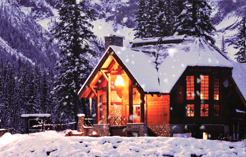 Video gif. It's snowing and we see a cozy cabin in the snow covered mountains with warm lights on.