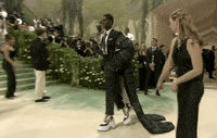 Met Gala 2024 gif. Alton Mason wearing a tweed ensemble and cape by Thom Browne, shuffles across the carpet confidently in towering platform boots, stopping to pop a leg and pose for the paparazzi.