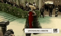 Met Gala 2024 gif. Gwendoline Christie serves expert poses with the statuesque posture of a Robert McGinnis figure, wearing a red Maison Margiela gown with a sheer black cape and bold, oversized blonde wig.
