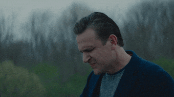 TV gif. Jason Segel in The Discovery frowns with his eyes closed as he puts his hand to his eyes. It is a dreary day with little color behind him and a gray sky. 