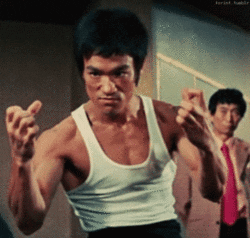 Movie gif. Martial artist Bruce Lee clenches his fists with an angry expression across his face. 