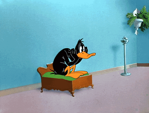Cartoon gif. Daffy Duck sits crunched on a tiny bed, head in his wings, thinking, worried.