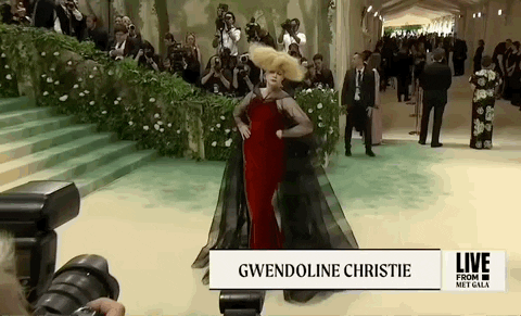 Met Gala 2024 gif. Gwendoline Christie serves expert poses with the statuesque posture of a Robert McGinnis figure, wearing a red Maison Margiela gown with a sheer black cape and bold, oversized blonde wig.