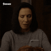 Video gif. A woman looks up from her phone, shaking her head, with a look of annoyance on her face.