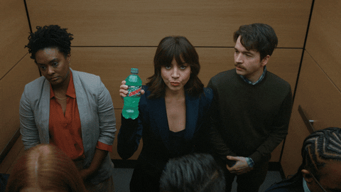 Sponsored gif. Aubrey Plaza is wearing a suit and stands in the middle of an elevator with office workers. Suddenly, the elevator lights blink, dust falls and the elevator rattles. Everyone around her freaks out but she stares at us deadpan as she holds a Mountain Dew Baja Blast in her right hand, cocking her head and saying, "Having a blast."