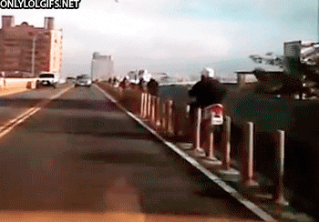 Video gif. Person rides a motorcycle on the side of a two-lane road, driving through a long line of barrier poles, knocking them over before falling off the bike and rolling onto the road.