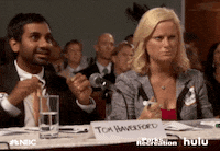 Parks and Recreation gif. Aziz Ansari as Tom dances in his seat next to Amy Poehler as Leslie who glares unhappily.