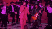 Oscars 2024 GIF. Ryan Gosling, dressed in a Barbie pink tuxedo and sunglasses, clutches the air with drama, flanked by Slash and a chorus line of dancing Kens.