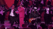 Oscars 2024 GIF. Ryan Gosling, dressed in a Barbie pink tuxedo and sunglasses, spins a background dancer Ken into his arms, who kisses him on the cheek, and spins out back into the chorus line.