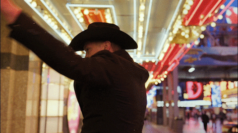 Celebrity gif. Jon Pardi, country singer, is walking through Las Vegas with a cowboy hat on and he has both hands in the air as he spins slowly and ecstatically.