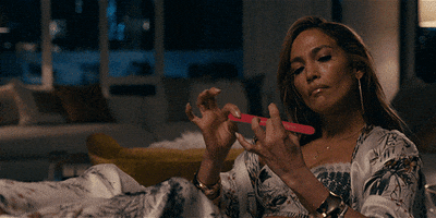 Movie gif. Reclining on a couch, Jennifer Lopez as Ramona in Hustlers purses her lips while filing her nails.