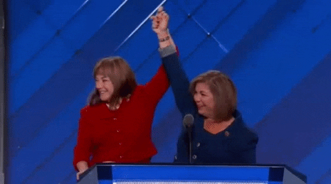 Politics gif. California representatives Loretta Sanchez and Linda Sanchez, wearing a red and a blue outfit respectively, hold hands, waving them in the air at the DNC 2016 podium.