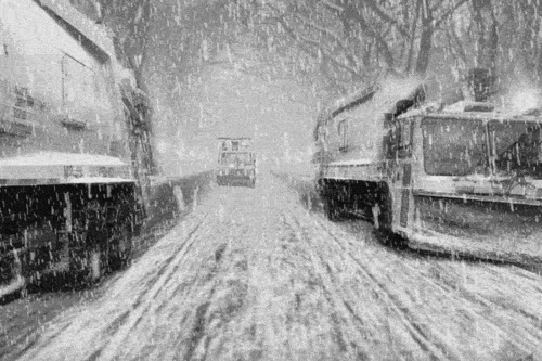 Video gif. Black and white footage of heavy snow falling on three snow plows lining a street that appears to move like a conveyor belt.