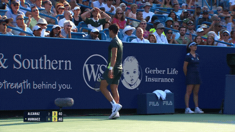 Sports gif. Clip of Carlos Alcaraz on a competition tennis court walking away from us towards the back wall of the court. He does two push ups like he's showing how strong he is, then turns towards us and smiles. 