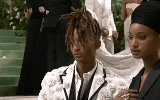 Met Gala 2024 gif. Closeup of Willow and Jaden Smith posing together. Willow is in a black blazer dress with hair styled like a crown and a black headwrap. Jaden is wearing an open dress shirt and white coat covered in flowers.