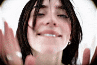 Music video gif. A scene from Billie Eilish's music video for "LUNCH" shows a close-up of Billie's face—who has long black hair—against a stark white background taken from a fisheye lens. She playfully bites her bottom lip and smiles while clapping for the camera.