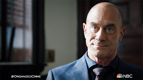 TV gif. Christopher Meloni as Detective Stabler on Law and Order cocks his head and smiles as if to say, “oh well.”