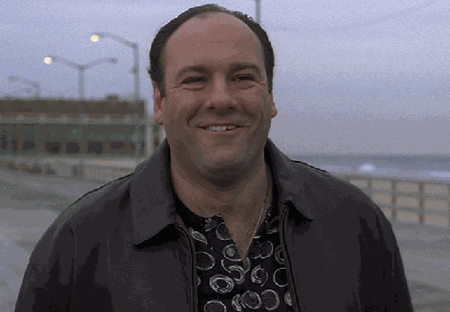 TV gif. James Gandolfini as Tony Soprano smiles and laughs to himself while walking along a pier.