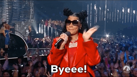 Video gif. Nicki Minaj at the 2023 VMAs, wearing dark oversized sunglasses and a red leather jacket with her hair in half-up-half-down high buns, holding a microphone while smiling and waving at the crowd. Text, "Byeeee!'