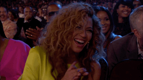 Celebrity gif. Beyonce's face lights up with laughter as she claps happily for someone at the BET Awards. She looks radiant with a neon yellow dress and curls that hang loosely over her face. Behind her, the rest of the crowd is also smiling and applauding.