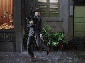Movie gif. Gene Kelly as Don Lockwood in Singin' in the Rain, holding a closed umbrella, happily tap dancing to and fro in the rain. 