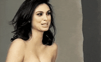 Celebrity gif. Morena Baccarin poses for a photoshoot without a shirt on. She smiles for the camera and a fan flows her hair gently.