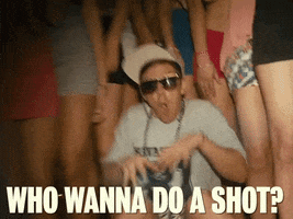 Music video gif. Jorma Taccone of The Lonely Island, dressed as a mid-00s rapper in oversized sunglasses and a crooked cap, crouched amongst women's legs, hands wide throwing battle gestures, raps at us "Who wanna do a shot?"