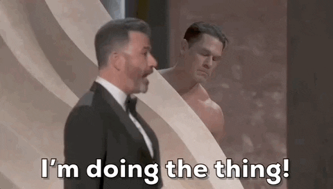 Oscars 2024 GIF. A potentially nude John Cena is behind a wall on stage and he is totally stressed out. Jimmy Kimmel stands in front of him and Cena gestures with his face for Kimmel to come to him now. Kimmel says, "I'm doing the thing!" but Cena insists. 