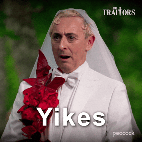 Sponsored gif. Alan Cumming as host of in The Traitors wearing a white suit with red flowers pinned to the left side and a white wedding veil grimaces and points. Text reads, “Yikes.”