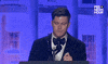 Video gif. Saturday Night Live's Colin Jost stands at a podium at the 2024 White House Correspondents' Dinner and delivers a joke. He says, "Wordle is here tonight" and pauses for a moment. He then closes his eyes in embarassment as he corrects himself and says "Sorry, I meant the New York Times."