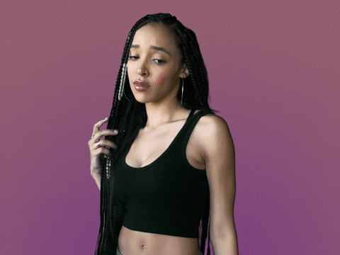 Celebrity gif. Tinashe twirls her waist-length hair, with braids in it, looking at us with a mild look of disgust, saying, "Ew."