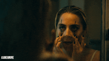 Movie gif. Lady Gaga as Harley Quinn in Joker wears thick black eyeliner as she stares in the mirror, using two fingers to pull at her lower eyelids. 