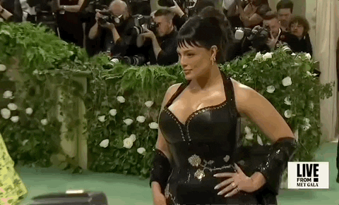 Met Gala 2024 gif. Ashley Graham wearing a custom Ludovic de Saint Sernin gown with black leather bodice, offers moody poses to the paparazzi as someone runs through frame behind her.