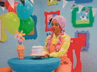 TV gif. Character in Happy Place wearing a lavender pastel wig, bright yellow checkered shirt, and coral-colored overalls sits at a table holding up a birthday cake in a brightly colored cartoonish set. She smiles sweetly and says, "Happy birthday bestie."