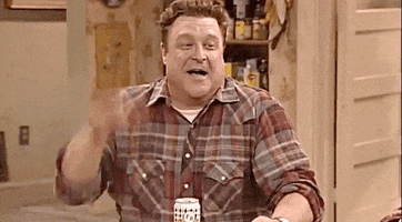 TV gif. John Goodman as Dan in Roseanne slaps the table and throws his head back laughing maniacally.