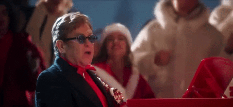 Celebrity gif. Elton John sits in front of a red piano, singing while dancers dressed in Santa Claus outfits dance happily behind him.