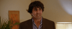Video gif. Jay Chandrasekhar in a brown suit raises his eyebrows and shrugs his shoulder as if to say, “Yeah well…”