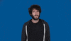Celebrity gif. Lil Dicky stands in front of a solid blue background, looking from side to side with a slightly confused frown. He puts a hand to his chest as he speaks. Text, "Who me? He grins."