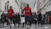 Ad gif. Two men wearing red soccer jerseys run down a city street, leading a crowd of people running with them and inviting more onlookers who join.