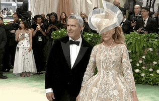 Met Gala 2024 gif. Sarah Jessica Parker poses with Andy Cohen. Parker is wearing a long sleeve scoop neck beige Richard Quinn sculptural corset gown with a bejeweled floral waist and cinched waistline paired with a gold and white Philip Treacy fascinator that sweeps in a wave-like pattern diagonally across her face. Cohen is wearing a classic black tuxedo smiling and nodding at the crowd.